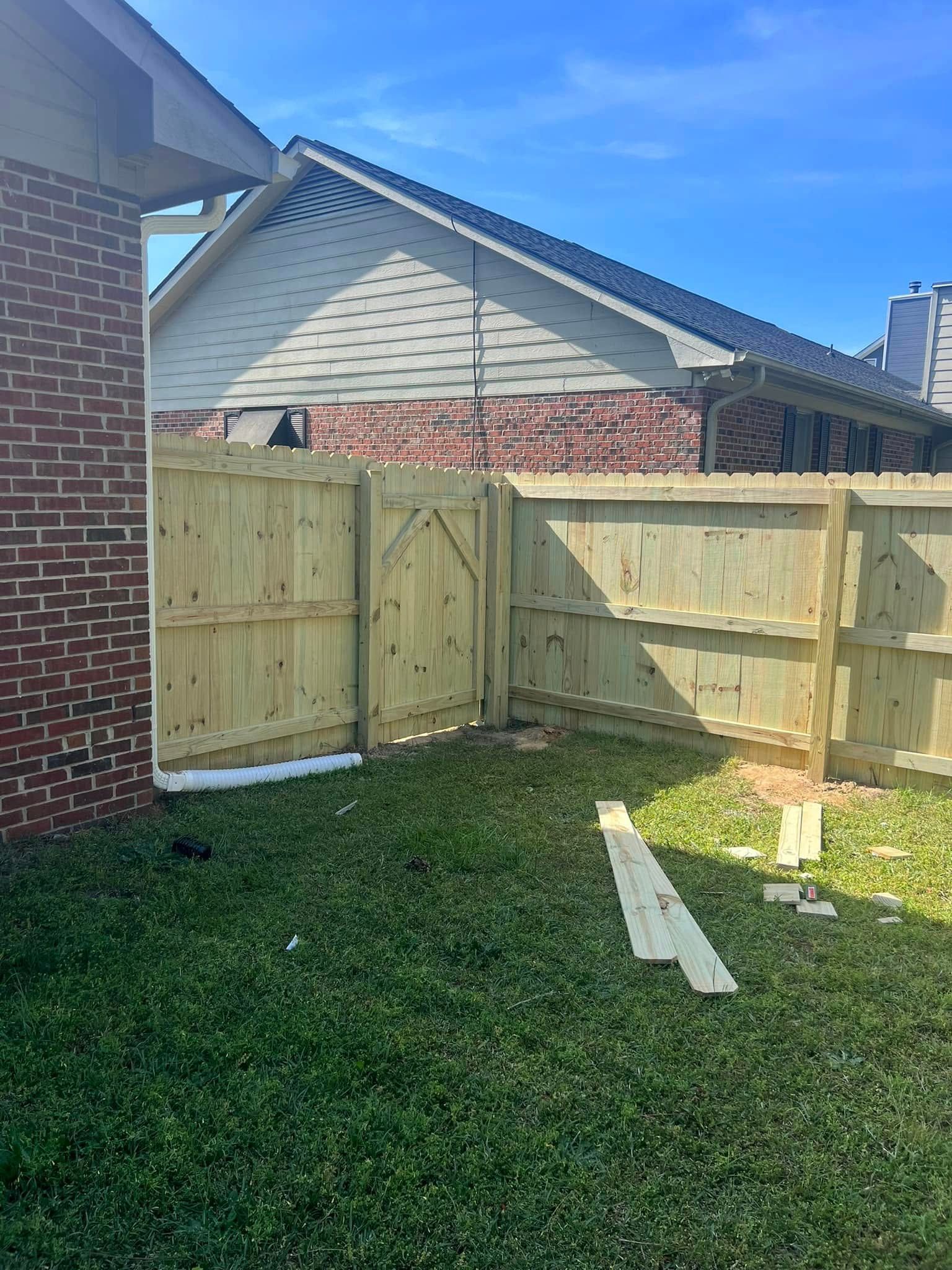 Newly built wooden fence in a grassy yard. Brick wall of a house is on the left, blue sky in the background.