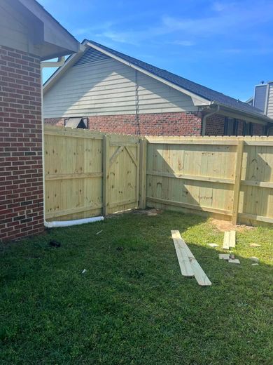 Wooden fence construction in a grassy backyard, next to a brick house, under a blue sky.