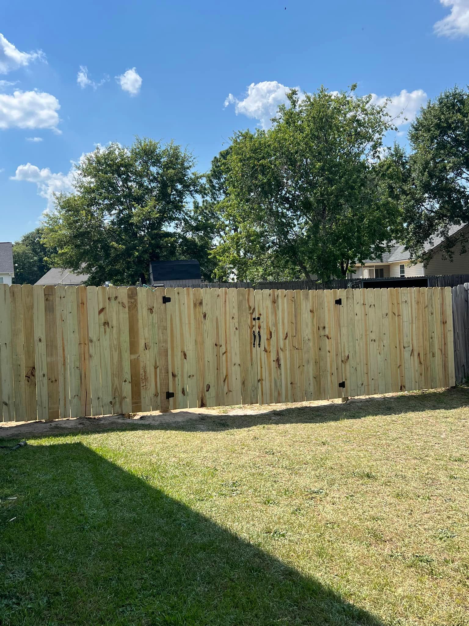 New wooden fence in a backyard with green grass and trees under a blue sky.
