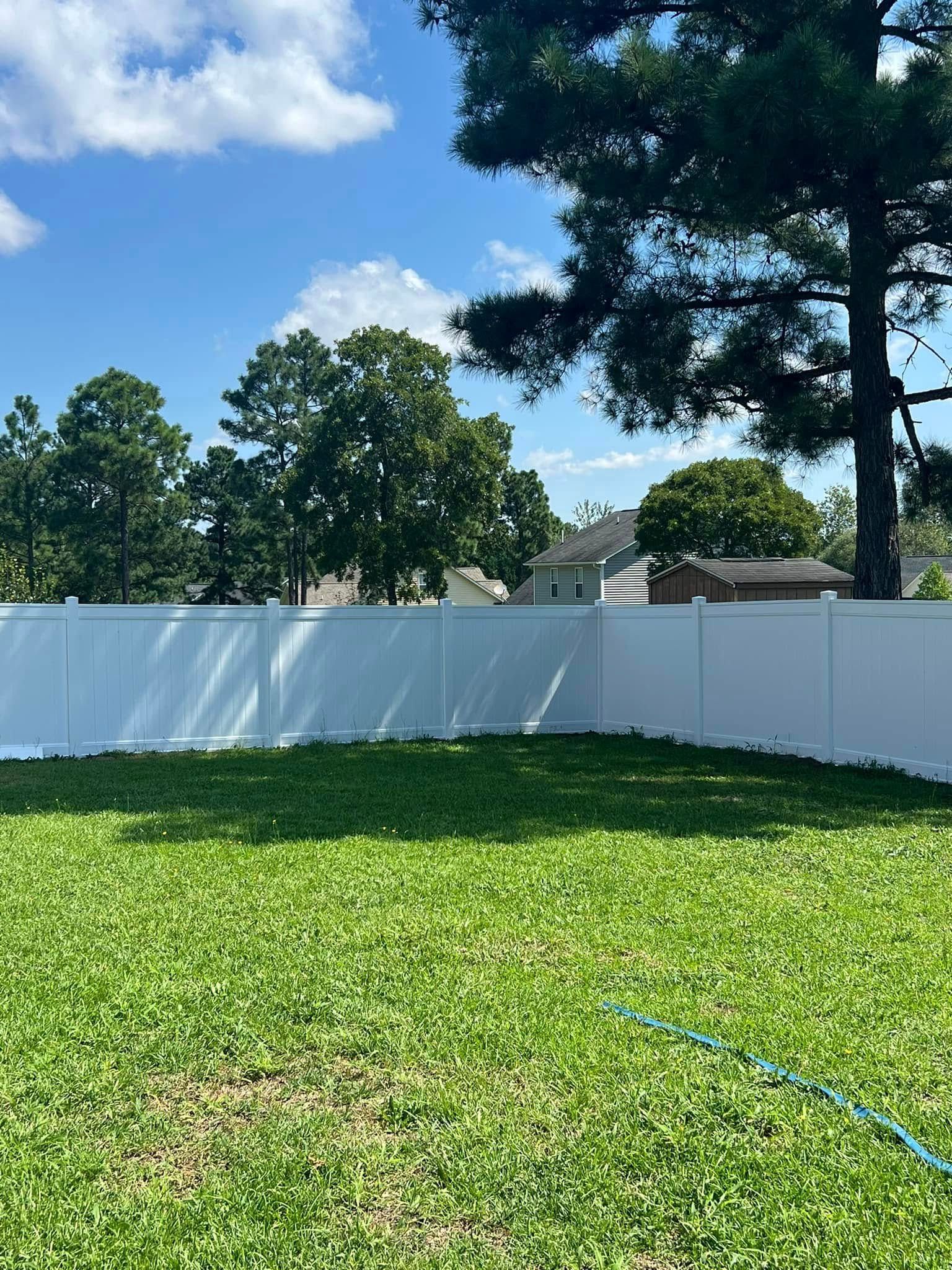 White fence encloses a grassy backyard under a blue sky, with trees and houses in the background.