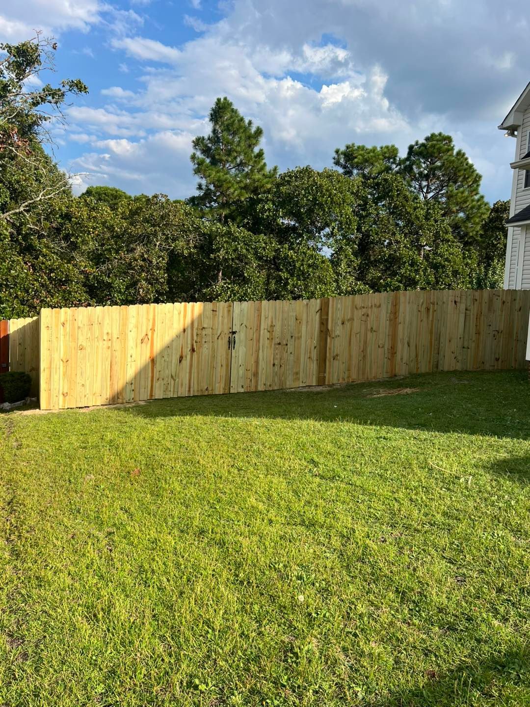 A wooden fence surrounds a grassy backyard under a partly cloudy blue sky.