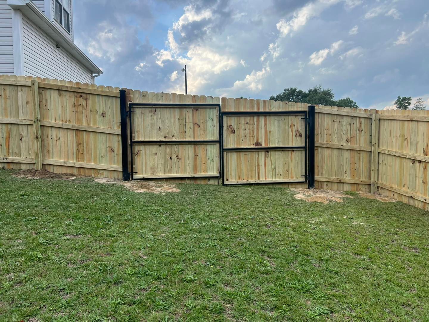 Wooden fence with black metal gate, in a grassy yard, under a cloudy sky.