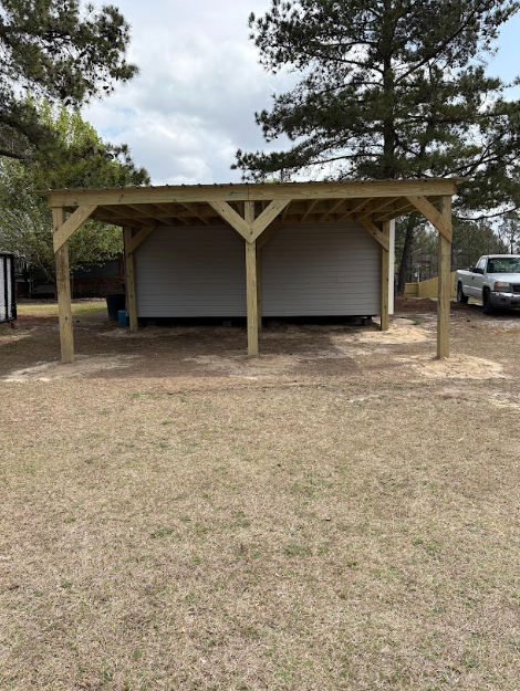 Wooden carport shelter over a white shed in a grassy outdoor area.