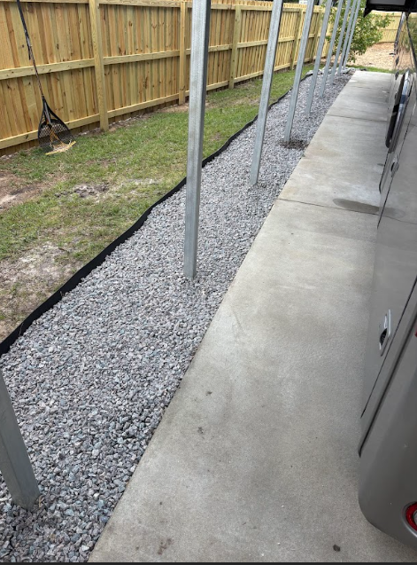 Gravel bed alongside a concrete walkway and wooden fence, with metal poles evenly spaced.