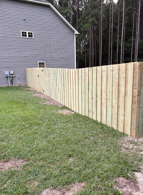 Wooden fence along a green lawn beside a gray house and a forest.