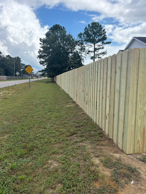A wooden fence runs along a grassy area beside a road, under a partly cloudy sky.