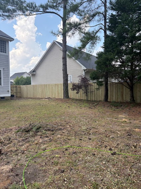 A backyard with a wooden fence, two houses, and trees. The ground is mostly dirt and some grass.