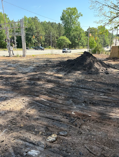 Muddy, dirt lot with a pile of dark soil, road, and trees in the background.