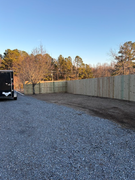 Gravel driveway with new wooden fence and trailer against a backdrop of trees under a blue sky.