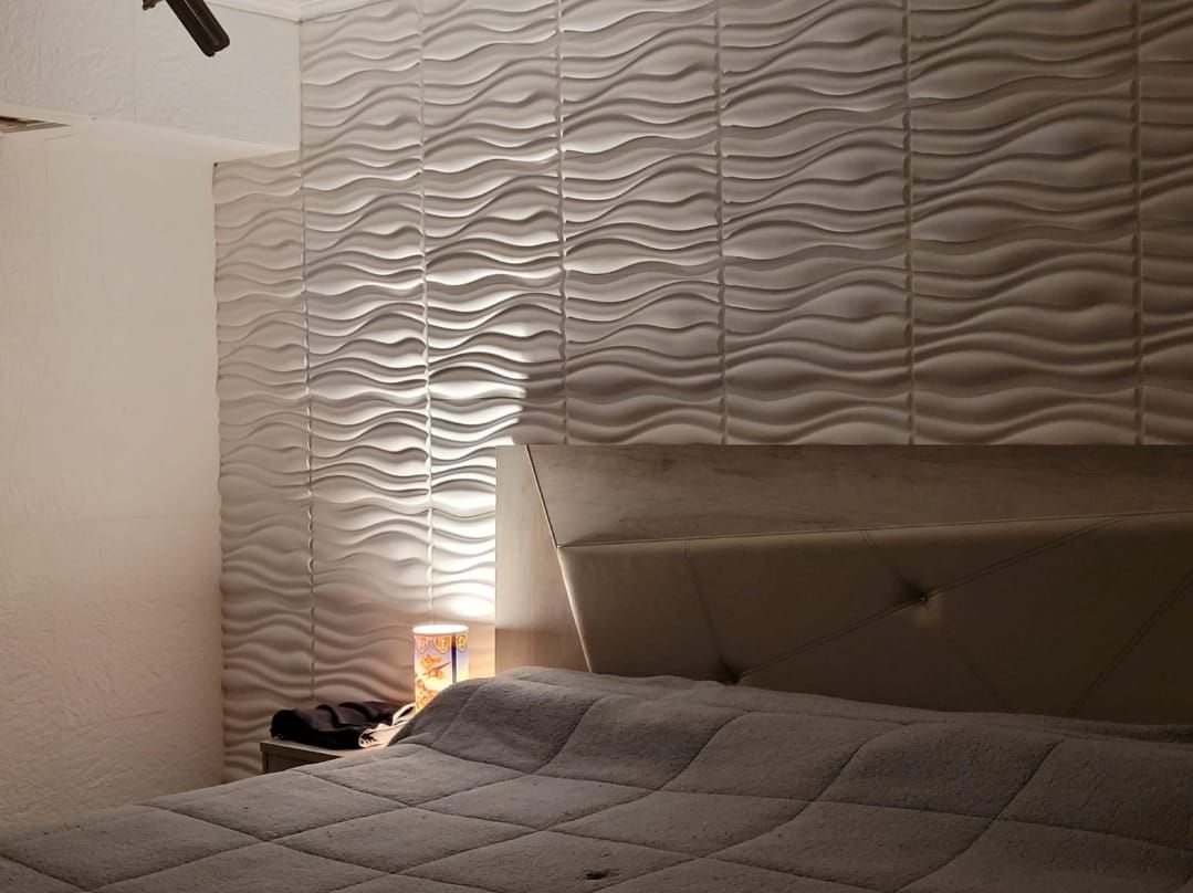 Dormitorio con paneles de pared blancos texturizados y ondulados, cama con cabecera capitoné y mesita de noche con lámpara.