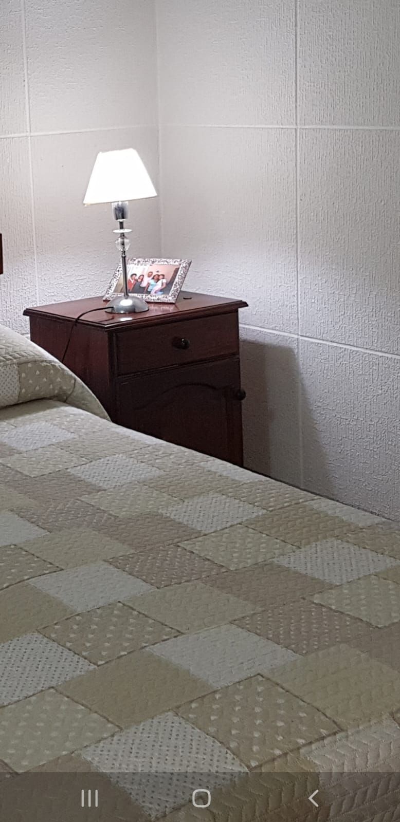 Dormitorio con mesita de noche de madera oscura, lámpara y cama con edredón estampado. Paredes blancas.
