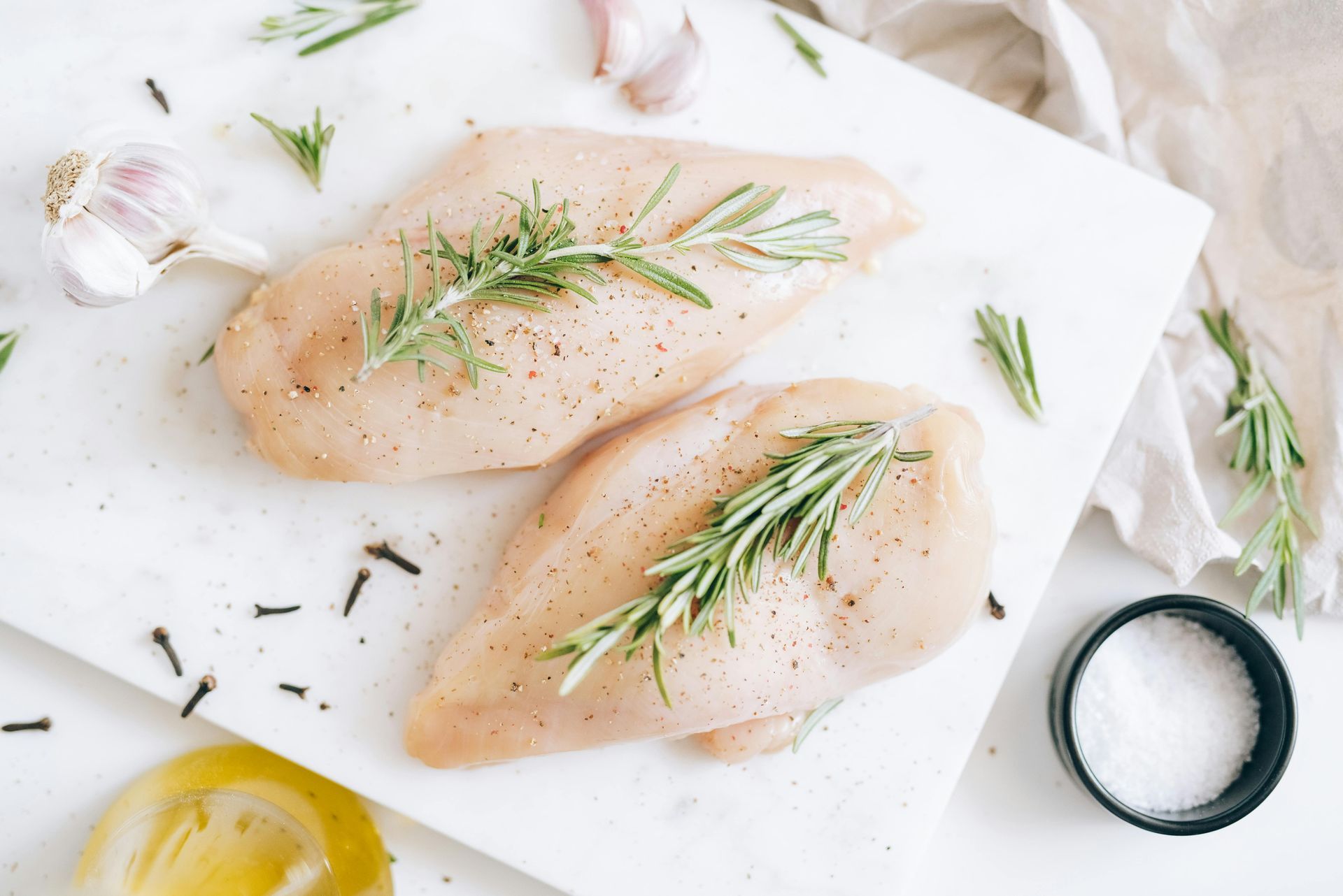 Two raw chicken breasts with rosemary on a cutting board.