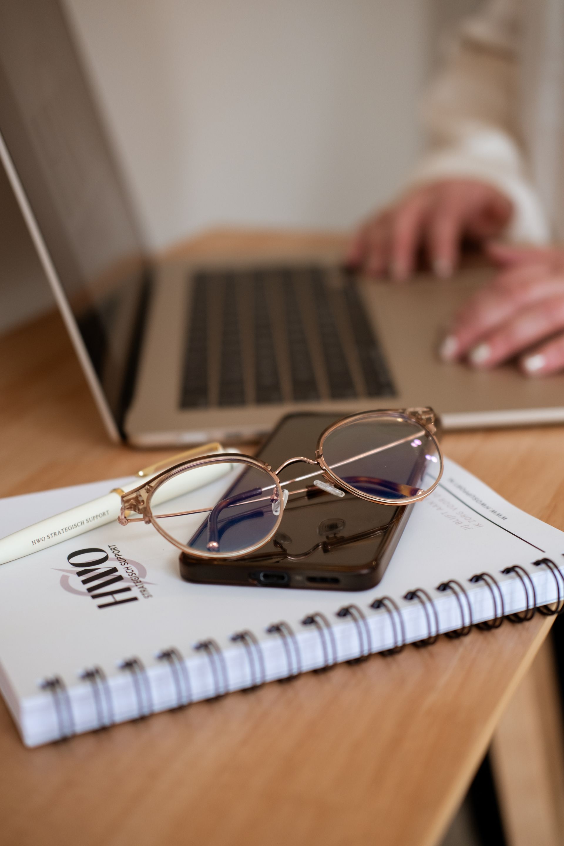 Vrouw in zwart pak aan een hoog houten bureau, schrijvend op een tablet. Beige muur, gedroogde bloemen in een vaas.