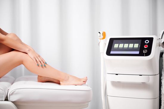 una donna è seduta su un letto con le gambe incrociate accanto a una macchina per la depilazione laser .