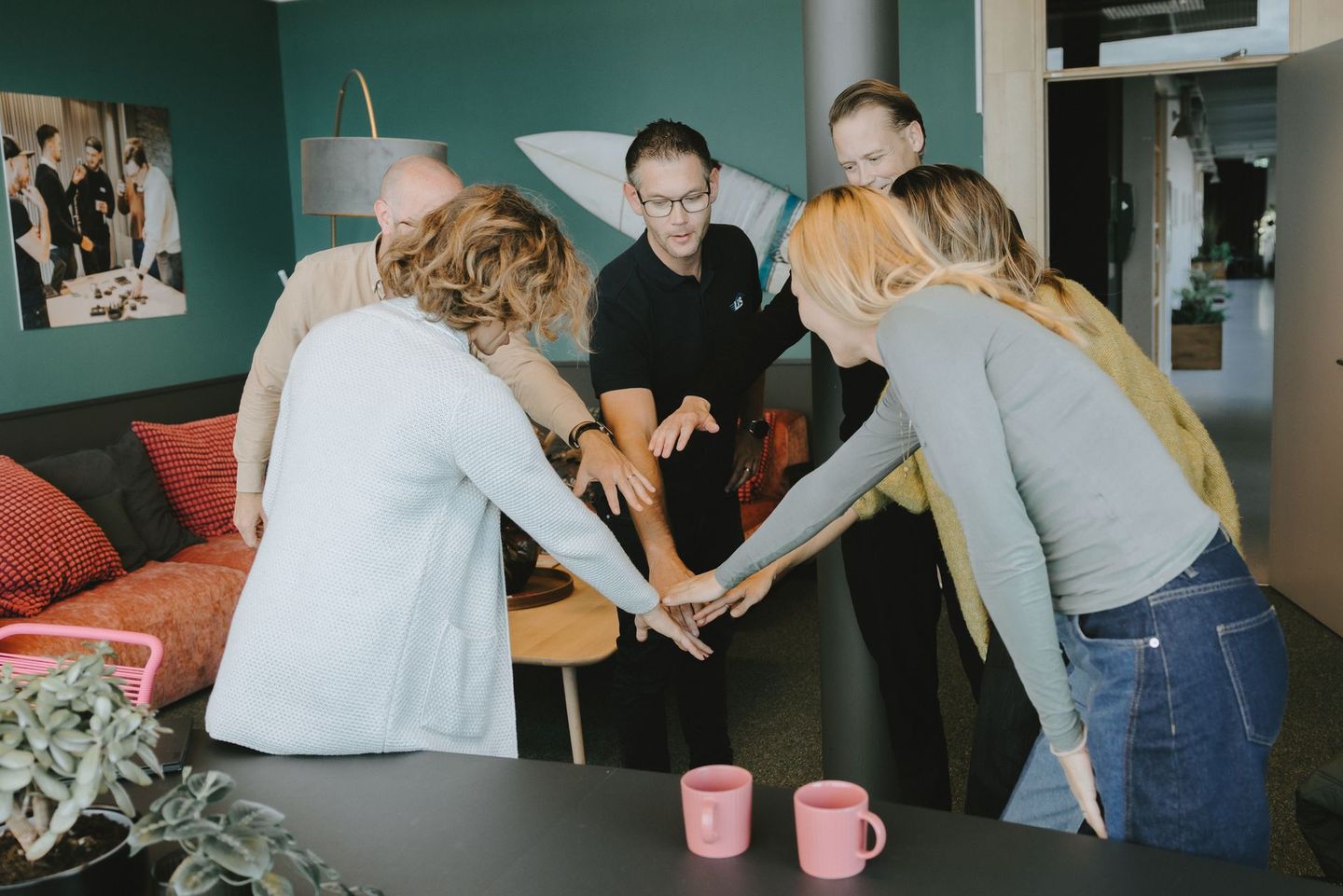 Grupp människor som håller händerna i en teamträff, inomhus med muggar på ett bord.