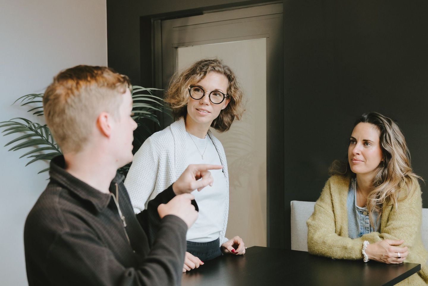 Tre personer i ett möte. Kvinna med glasögon pekar på personen till vänster. Mörkt rum, svart bord.