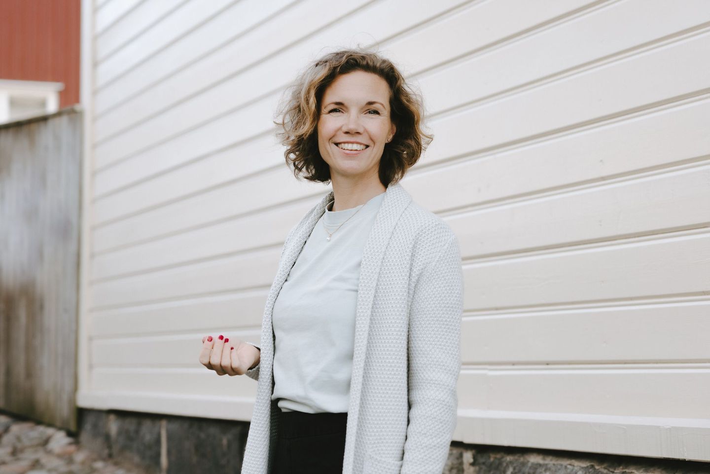 Kvinnan ler, poserar utomhus, klädd i en grå kofta och ljusgrön topp, mot en vitpanelerad vägg.