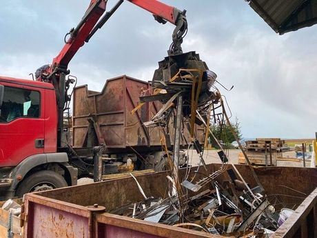 Red truck crane loading scrap metal into a dumpster under a cloudy sky.