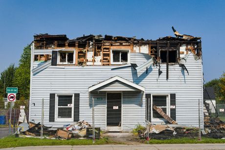 House damaged by fire, roof collapsed, windows charred. Debris in front on a sunny day.