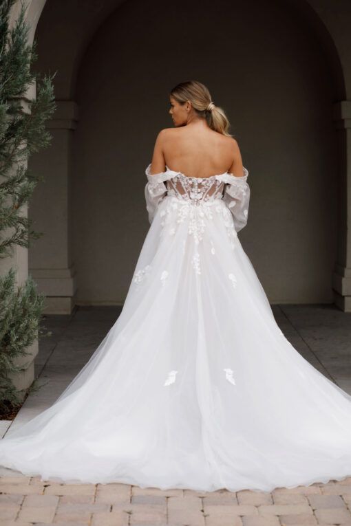 Lady stood in an Ivory/Rum wedding dress by Martina Liana. The wedding dress has a floaty tulle overlay with floral lace motifs and a rum lining. It also has a sheer illusion tulle back with a zip and buttons.