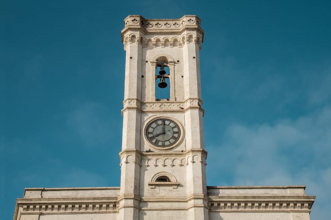 Torre dell'orologio in pietra bianca con campanile aperto sullo sfondo di un cielo azzurro limpido.