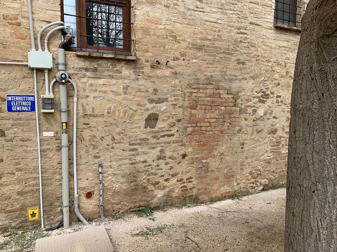 Sulla destra si vedono il muro di un edificio in pietra con una canalina elettrica, un cartello blu, una piccola finestra e un tronco d'albero.