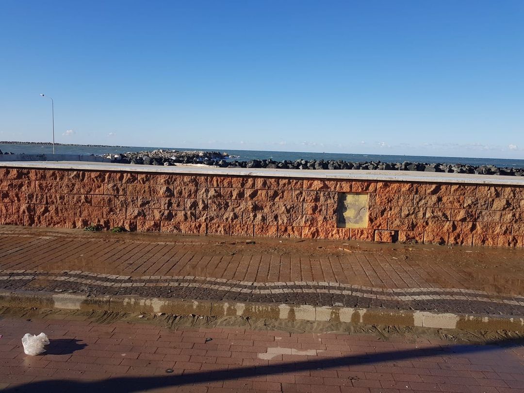 Un basso muro di pietra di colore bruno-rossastro delimita una passeggiata lastricata che si affaccia su un mare calmo sotto un cielo azzurro e limpido.