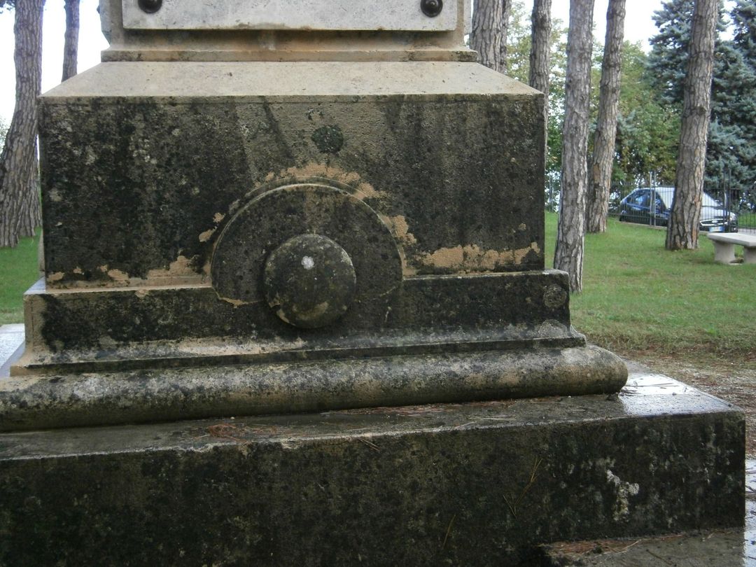 Un piedistallo in pietra consumata dal tempo con un rilievo decorativo circolare, situato in un parco all'aperto con alberi sullo sfondo.