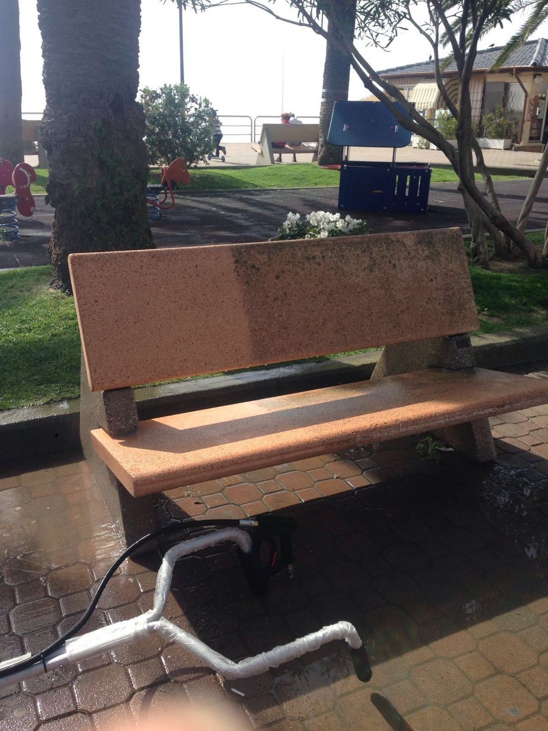 Un'idropulitrice pulisce metà di una panchina di pietra in un parco, evidenziando il netto contrasto tra la superficie pulita e quella sporca.