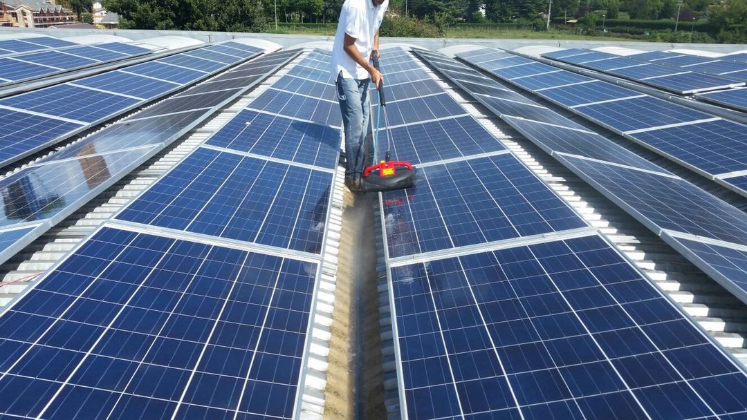 Una persona pulisce lunghe file di pannelli solari su un tetto utilizzando una spazzola rotante specializzata.
