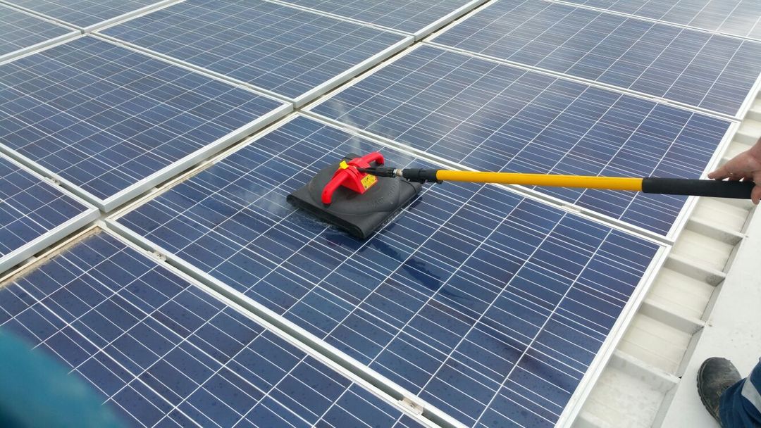 Un operaio utilizza una spazzola a manico lungo, rossa e nera, per pulire una fila di pannelli solari su un tetto.