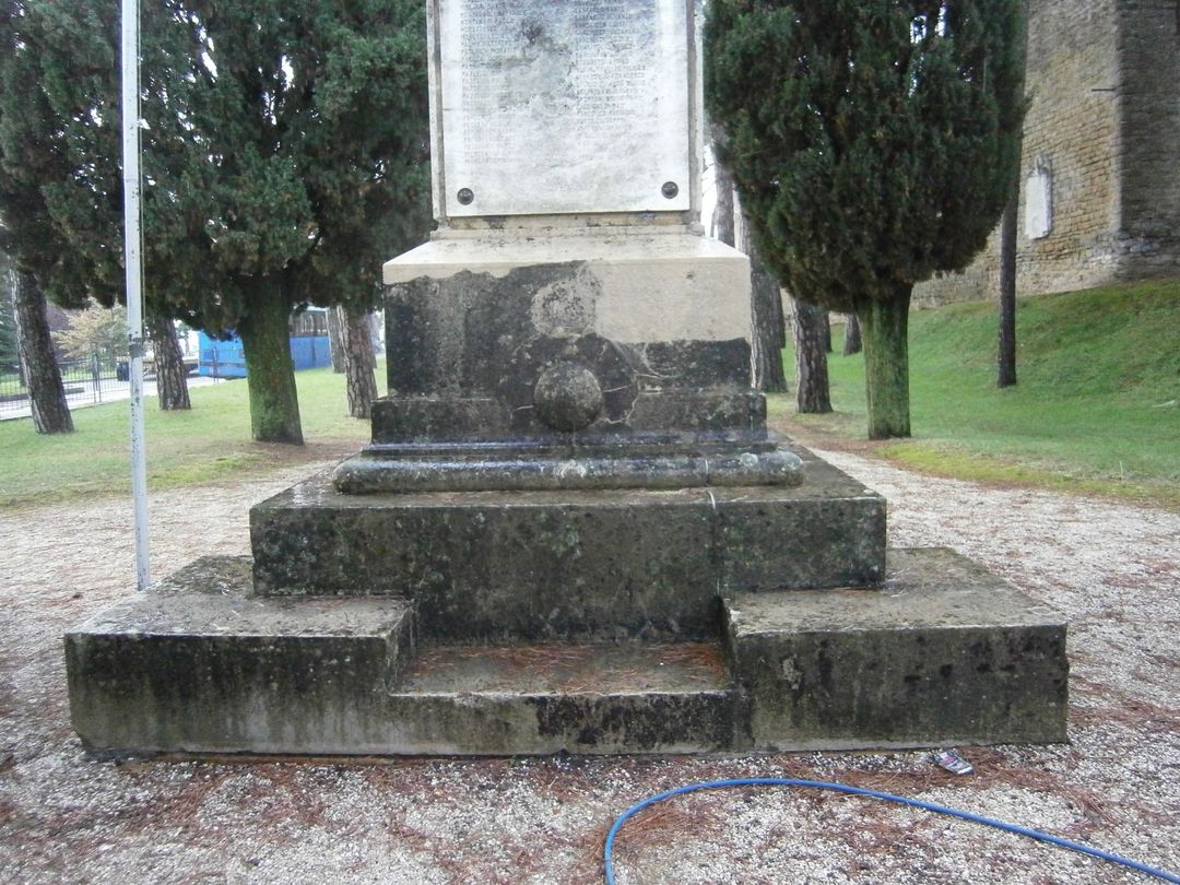 Una base monumentale in pietra con macchie scure dovute agli agenti atmosferici, posta su una superficie di ghiaia all'aperto con cipressi sullo sfondo.