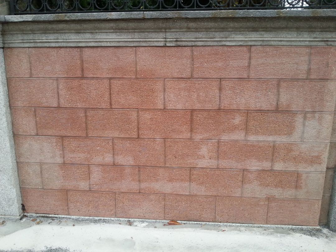 Un muro formato da blocchi rettangolari di pietra di colore rosa-rossastro, con una cornice superiore in pietra grigia e una recinzione metallica sovrastante.