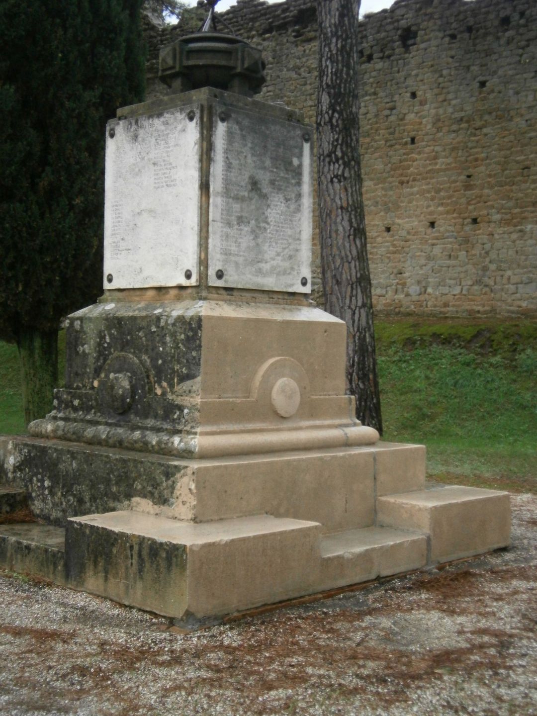 Un monumento in pietra consumata dal tempo, con una targa centrale in marmo, si erge all'aperto di fronte a un muro di pietra e ad alcuni alberi.