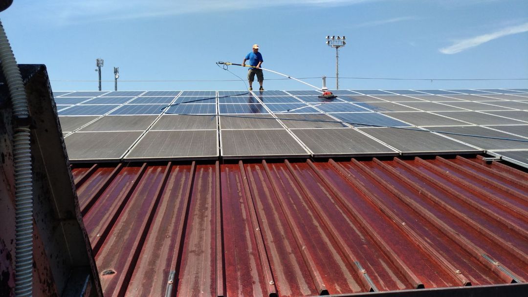 Sotto un cielo azzurro, una persona utilizza una spazzola dal manico lungo per pulire i pannelli solari installati su un tetto di lamiera ondulata rossa.