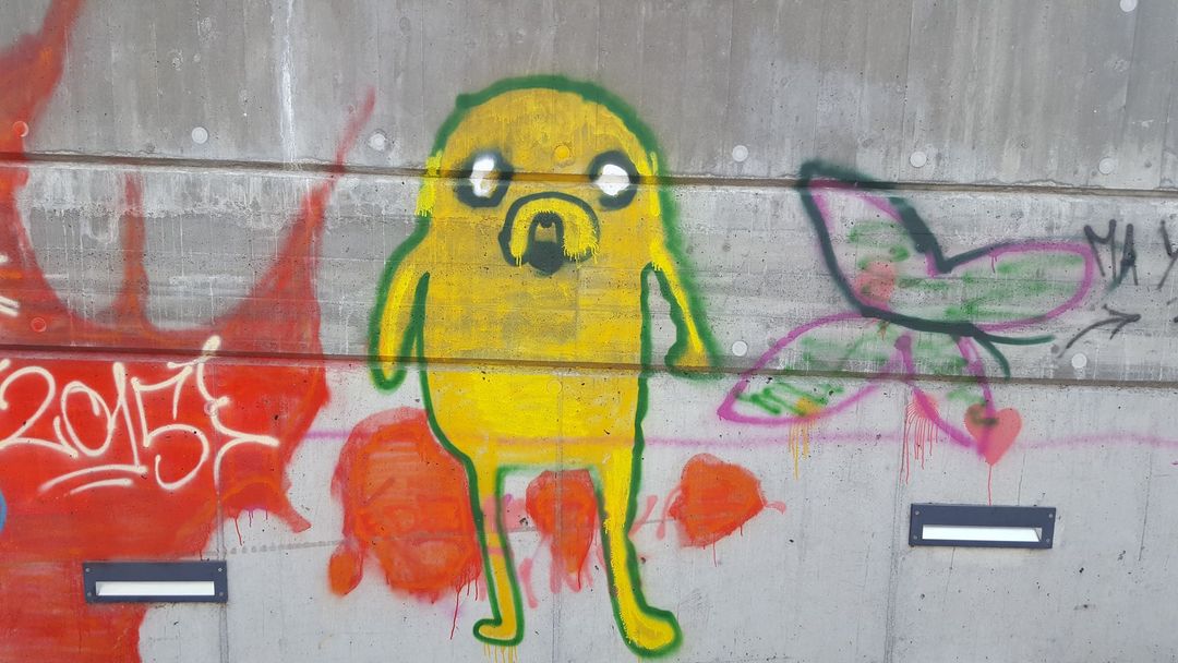 Un graffito realizzato con vernice spray raffigurante il personaggio dei cartoni animati giallo Jake il cane è dipinto su un muro di cemento, accanto a una farfalla rosa dipinta.