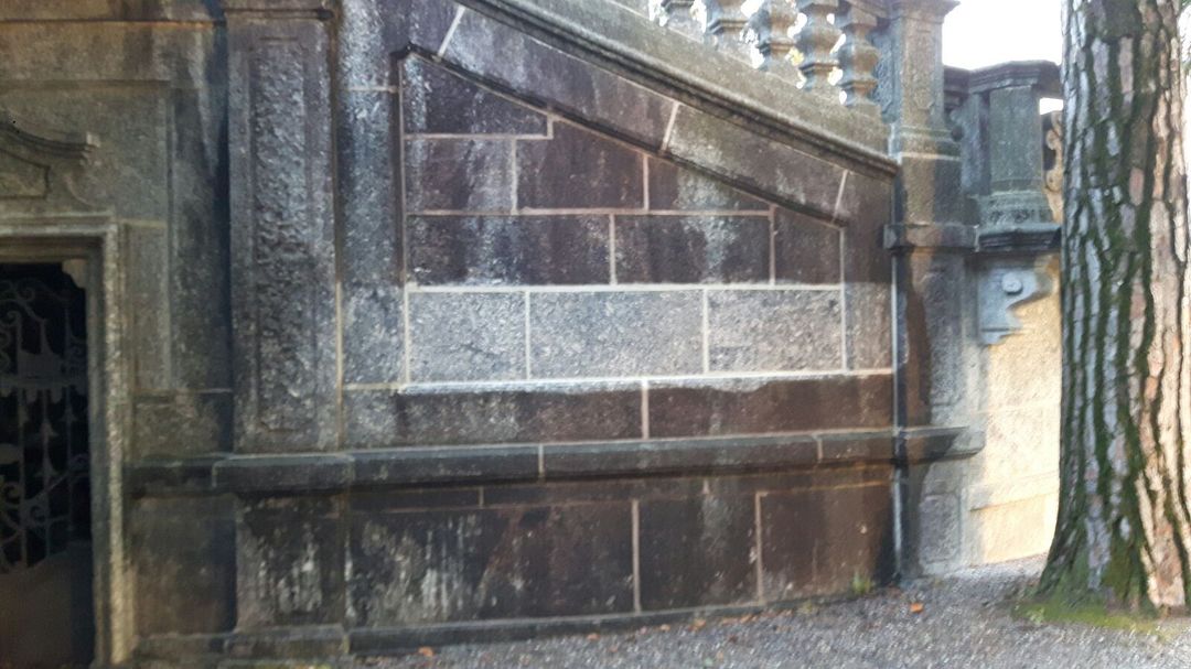 Una scalinata in pietra con blocchi rettangolari chiari e scuri, accanto a una porta in ferro battuto decorata e a un albero.