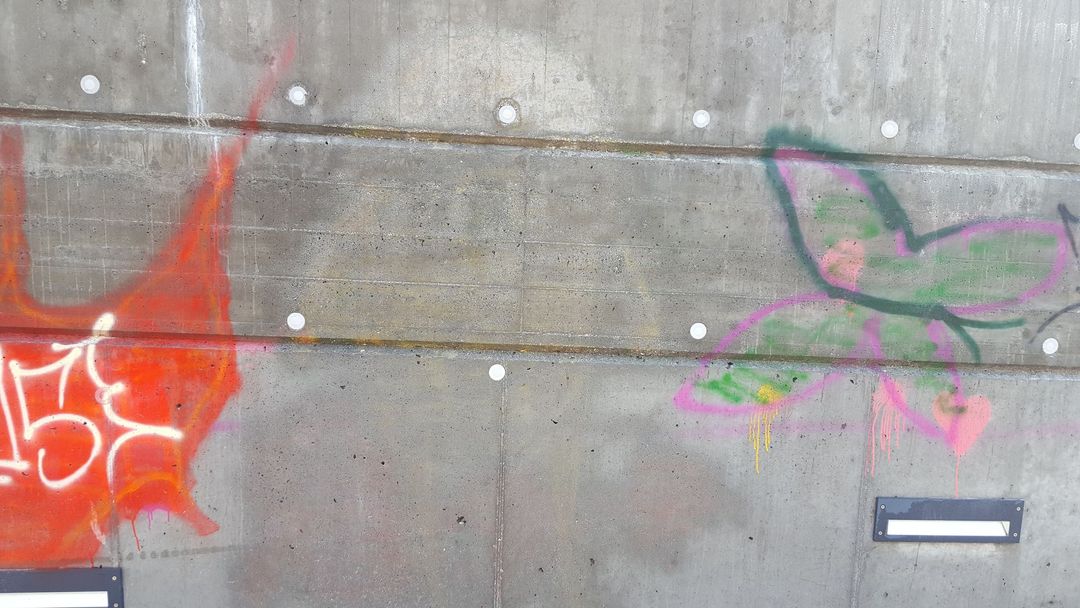 Un muro di cemento grigio con giunture orizzontali, su cui spiccano un graffito arancione acceso sulla sinistra e una farfalla dipinta.