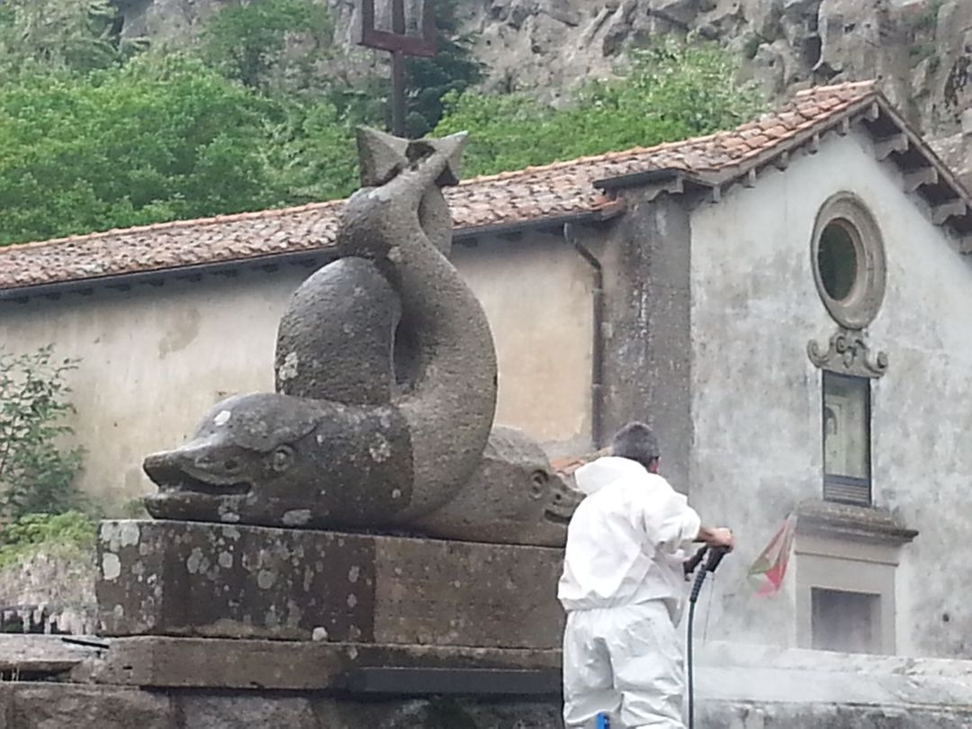 Un operaio con indosso una tuta protettiva bianca pulisce una scultura in pietra a forma di delfino davanti a un edificio rustico con il tetto di tegole.