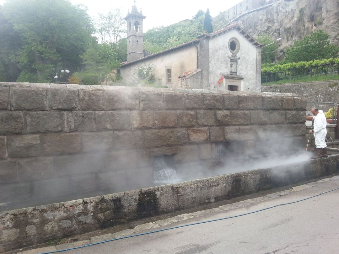 Una persona con indumenti protettivi pulisce con acqua calda e vapore una fontana di pietra vicino a una chiesa in pietra.