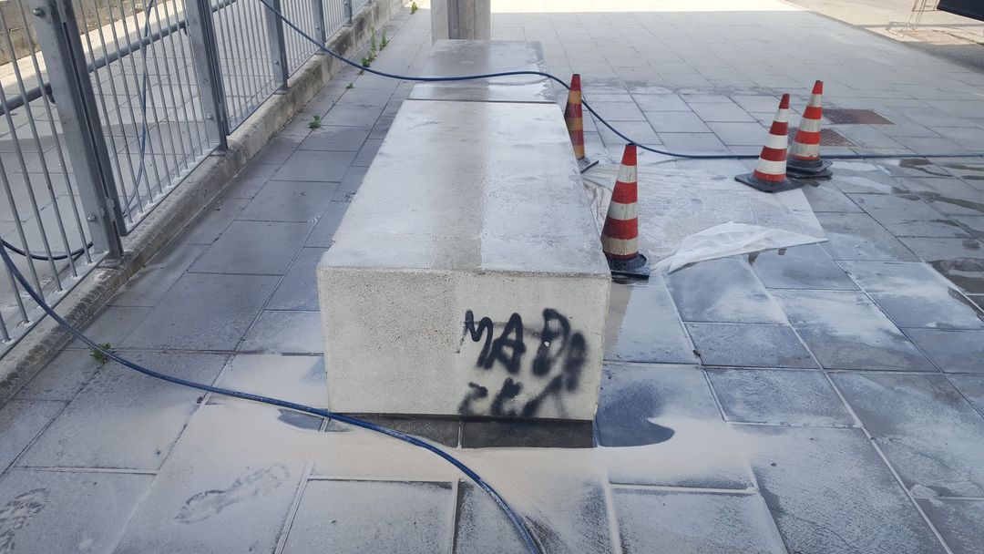 Una lunga panchina rettangolare in pietra con graffiti neri, circondata da tre coni stradali su una superficie urbana piastrellata.