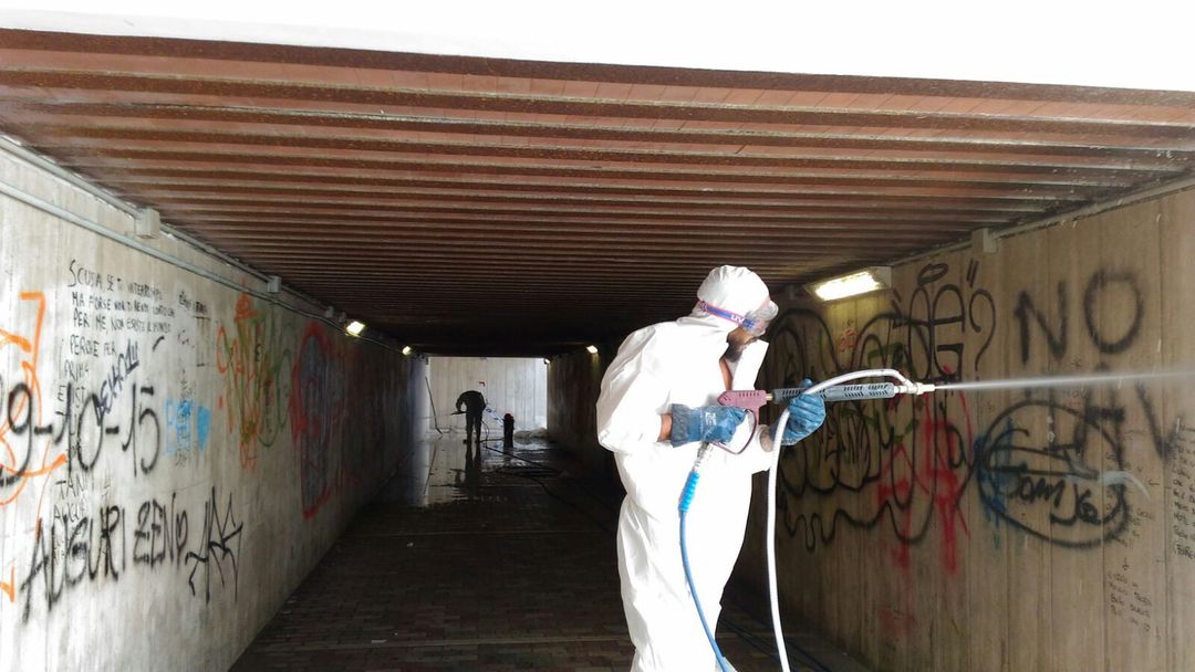 Una persona con indosso una tuta protettiva bianca rimuove i graffiti dalle pareti di un sottopassaggio in cemento con un'idropulitrice.