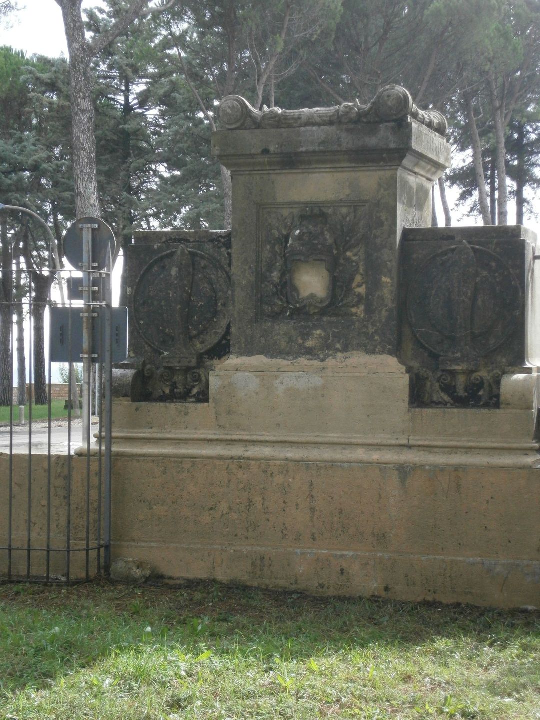 Un monumento in pietra con pannelli decorativi scolpiti, sullo sfondo di alberi e una recinzione metallica.