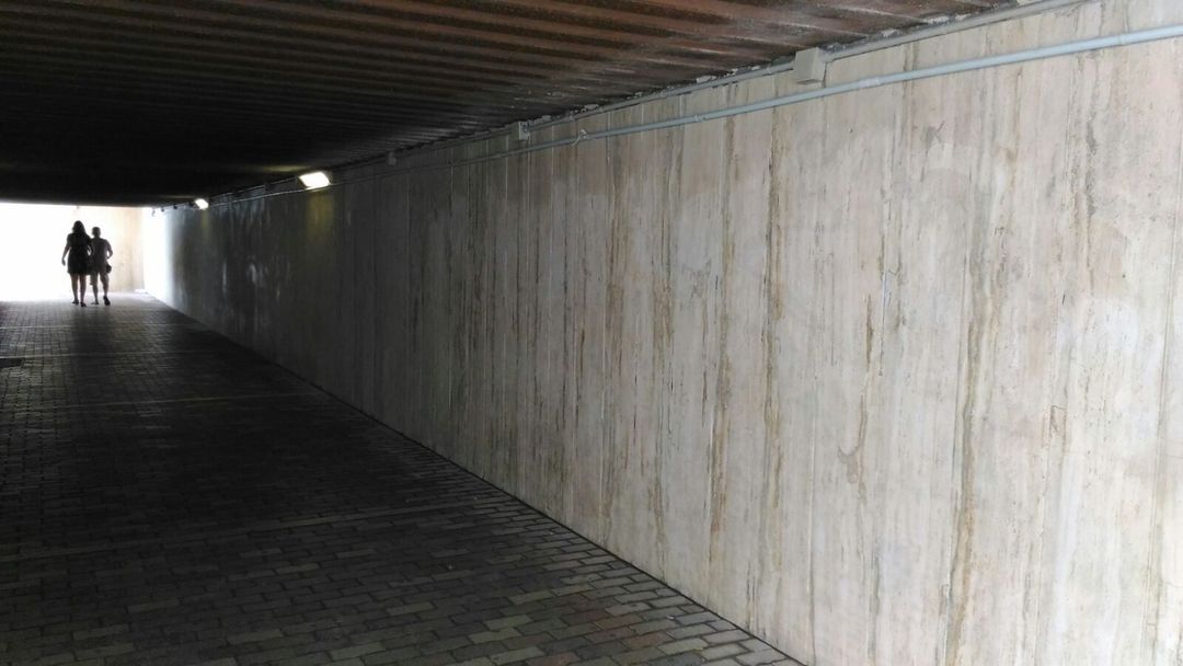 Un tunnel pedonale scarsamente illuminato, con pareti in pietra chiara e pavimento lastricato, da cui si intravedono due sagome in lontananza.