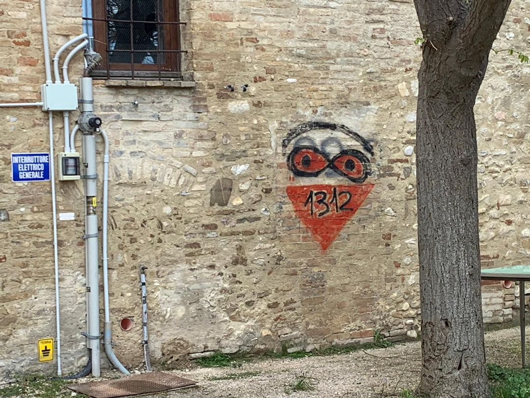 Su un muro di pietra è presente un graffito con un volto stilizzato sopra un triangolo arancione contenente il numero 