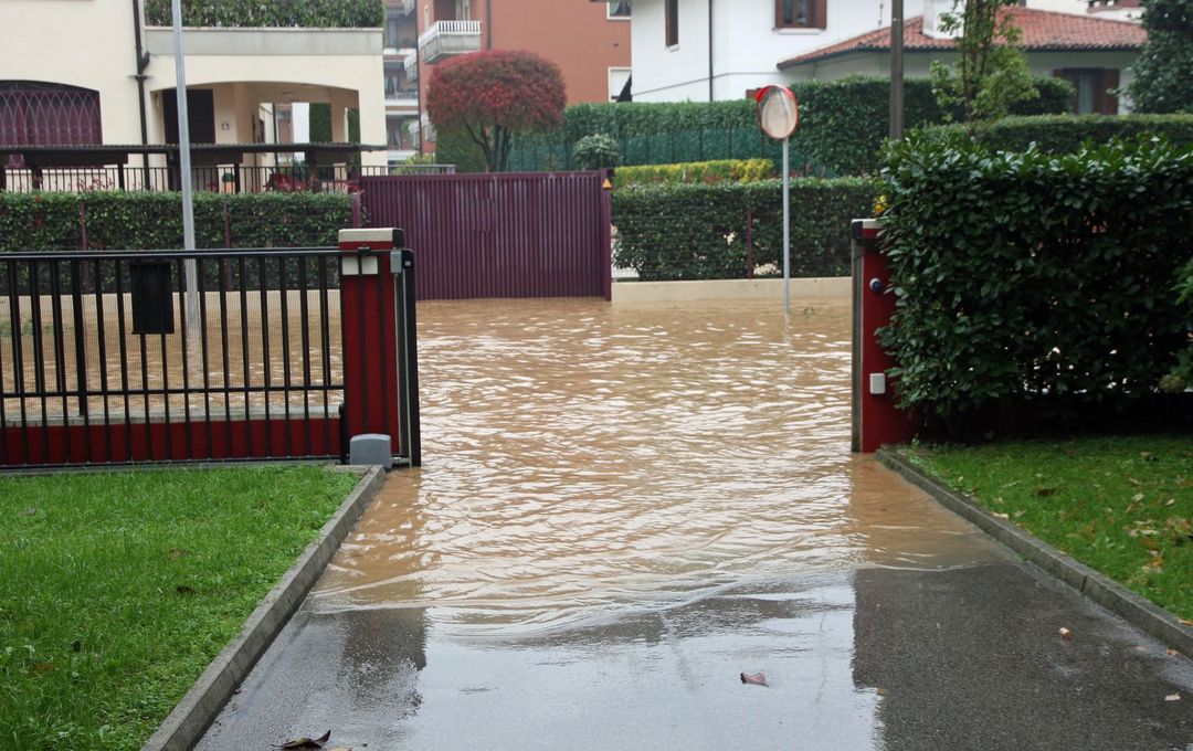 Un vialetto d'accesso allagato e una strada residenziale con acqua torbida e marrone che ricopre il marciapiede tra due cancelli.
