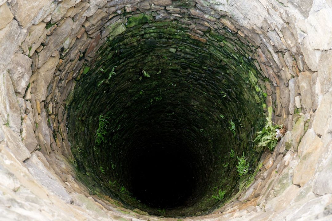 Un pozzo cilindrico rivestito di pietra che si affaccia su un abisso oscuro e profondo, con muschio che cresce sulle pareti interne.