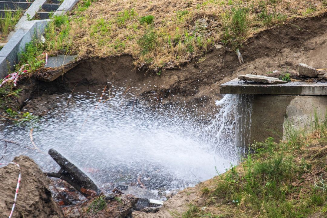 L'acqua schizza violentemente da un tubo rotto vicino a una struttura in cemento, erodendo la scarpata di terra di una collina.