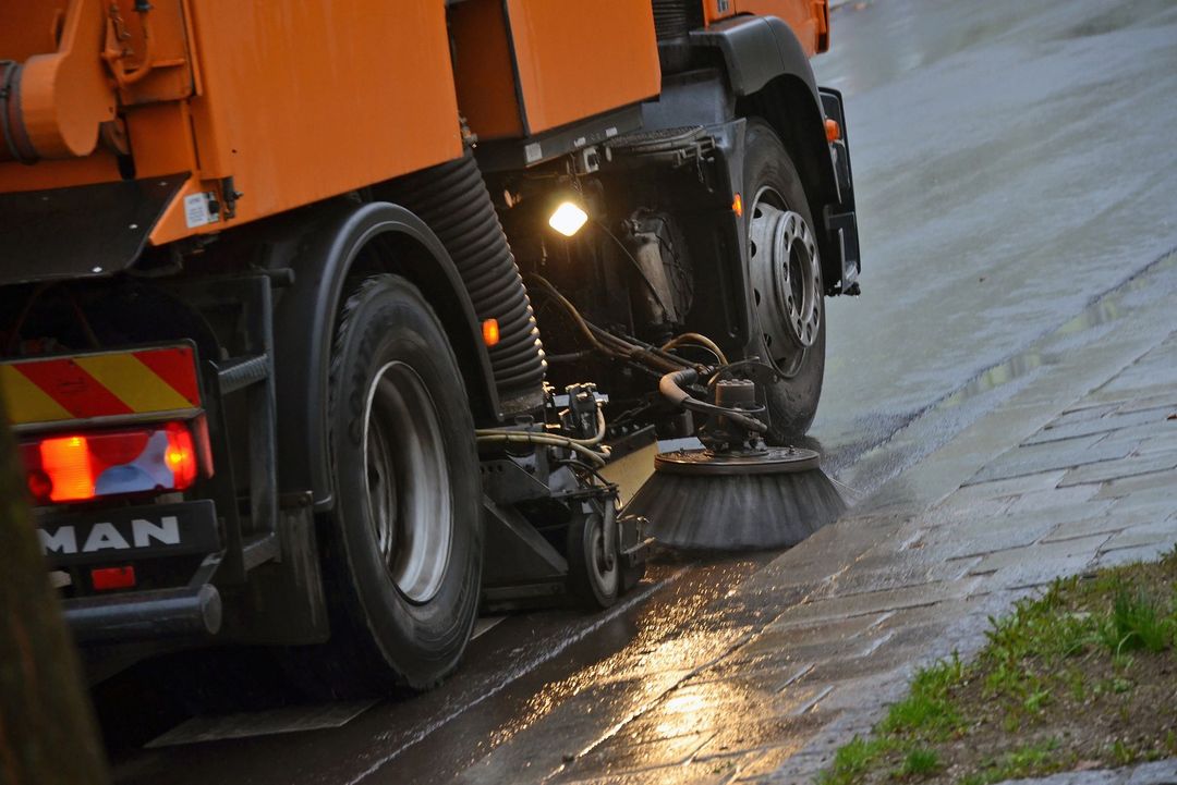 Un camion spazzatrice arancione pulisce una superficie stradale umida e bagnata con la sua spazzola circolare rotante.