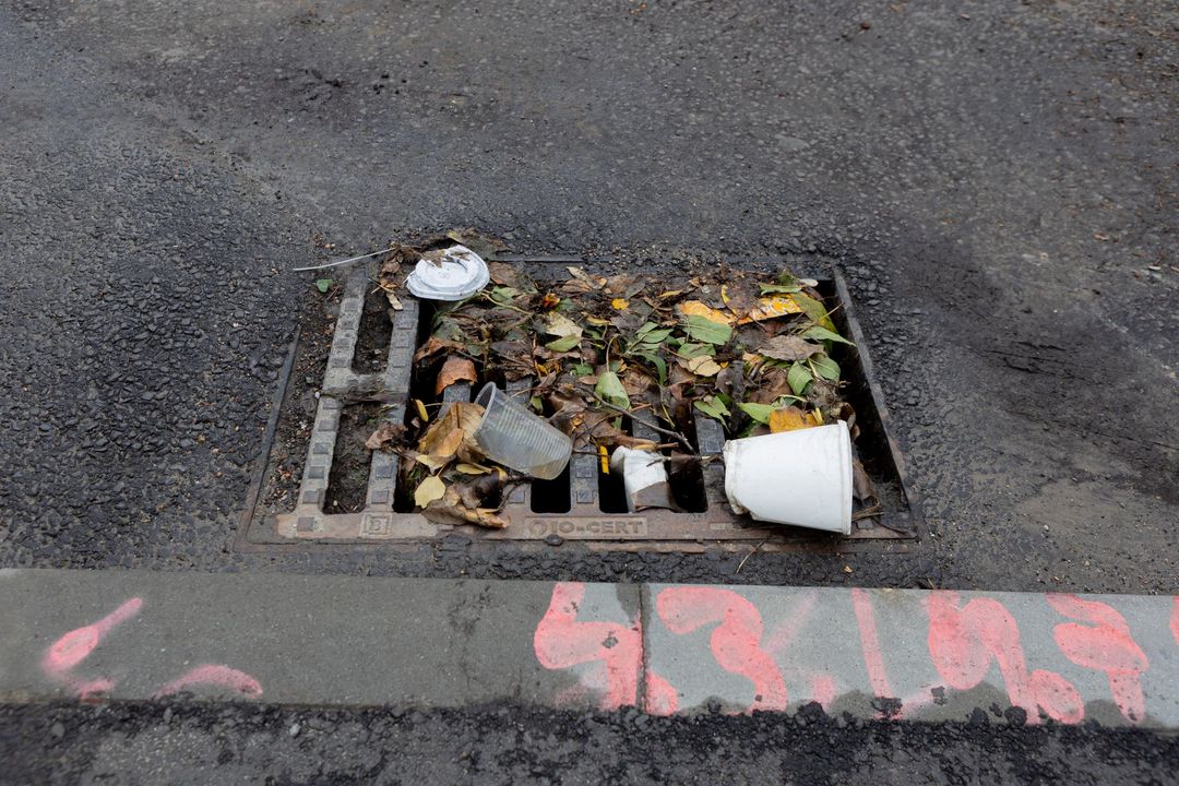 Un tombino ostruito all'angolo di una strada, pieno di foglie cadute e bicchieri di plastica bianca abbandonati.